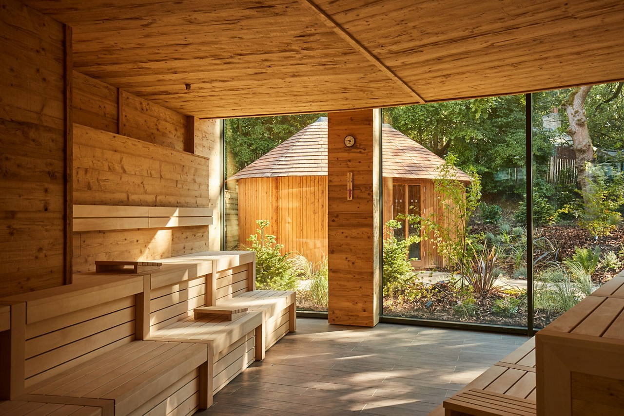 Sauna benches line a wood-clad room, basking in sunlight streaming through floor-to-ceiling windows; outside, a small round-roof wooden hut sits in a landscaped garden with trees and shrubs.